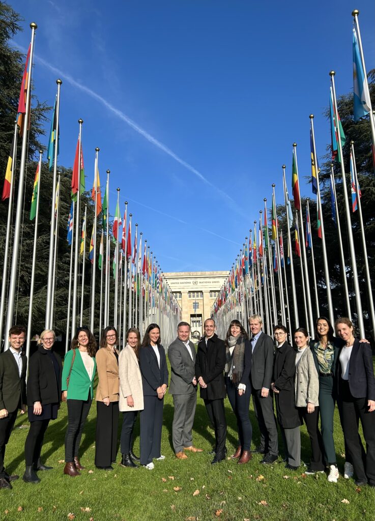 A group of people standing in front of the United Nations flags in Geneva, under a clear blue sky on a sunny day